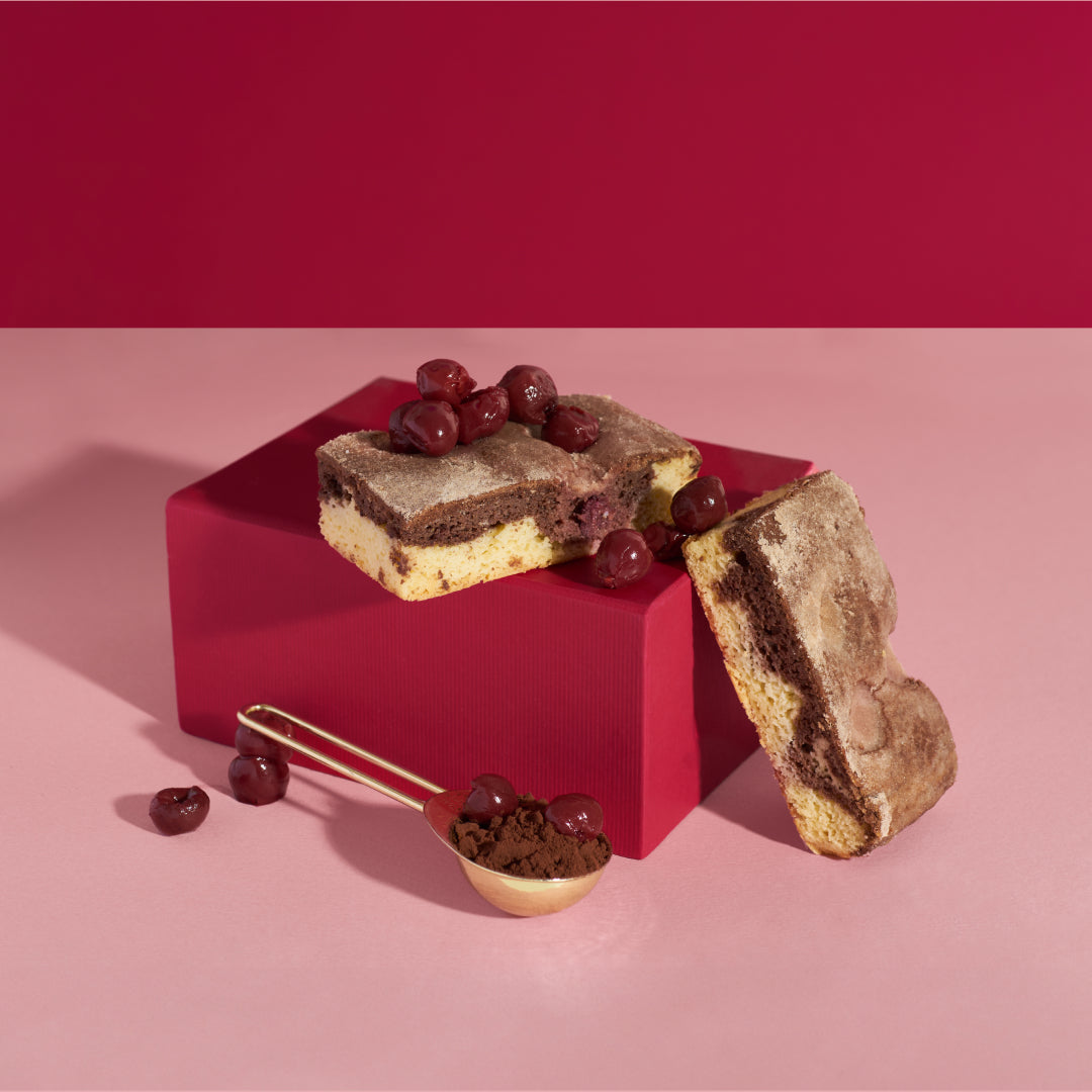 Marmor-Sauerkirsch Kuchen drapiert auf rosafarbenem Block mit Sauerkirschen und einem Löffel Kakao daneben